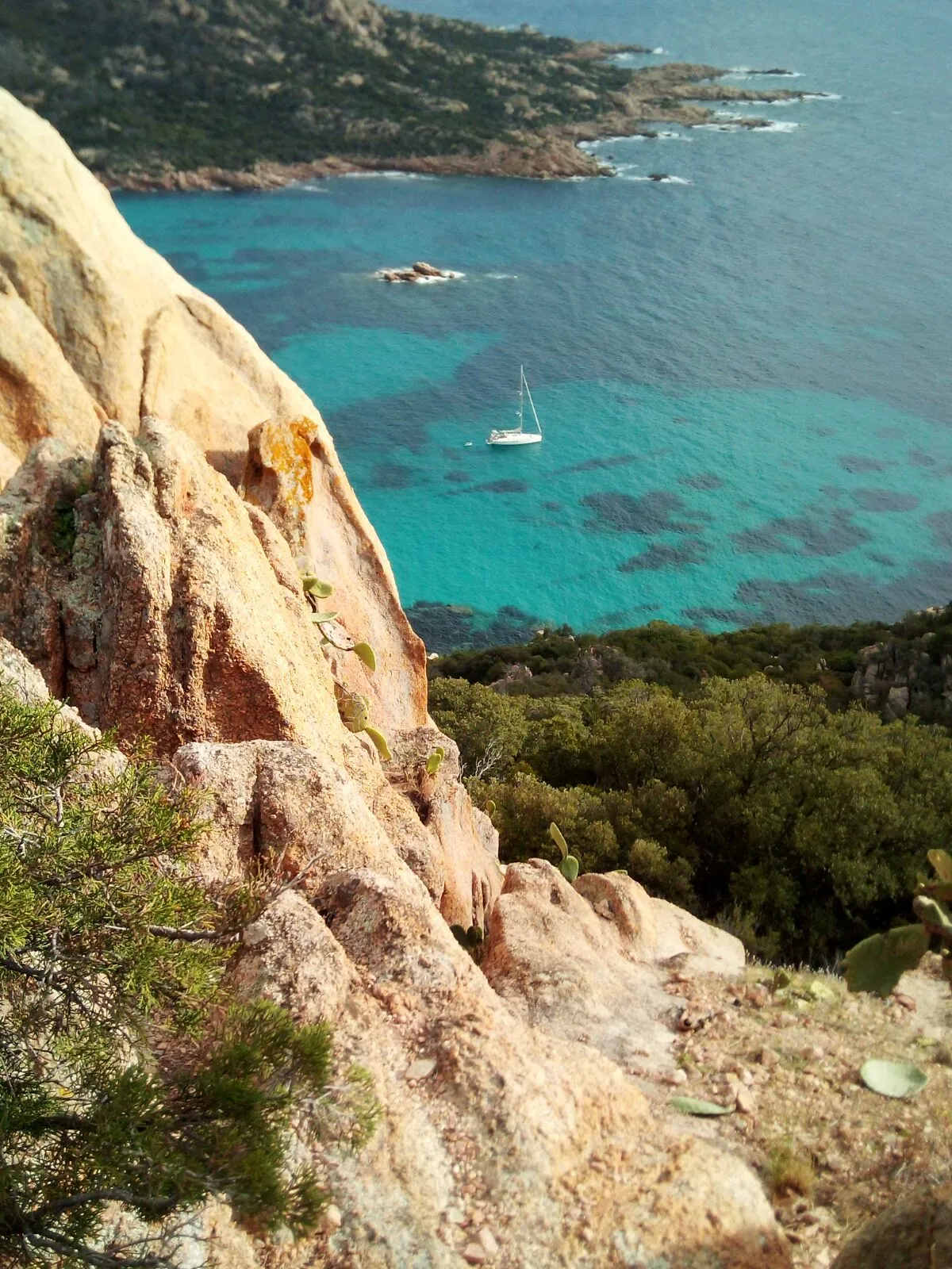 Vue plongeante depuis les falaises rocheuses de Corse-du-Sud sur un voilier au mouillage dans une eau turquoise.