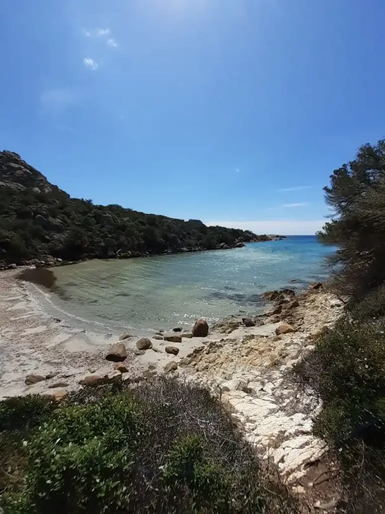 Vue sur une plage de sable isolée bordée de maquis dense en Corse. Des rochers de granite sont éparpillés sur le rivage et dans l'eau bleu clair, sous un soleil radieux. Un chemin de terre est visible sur le côté droit.