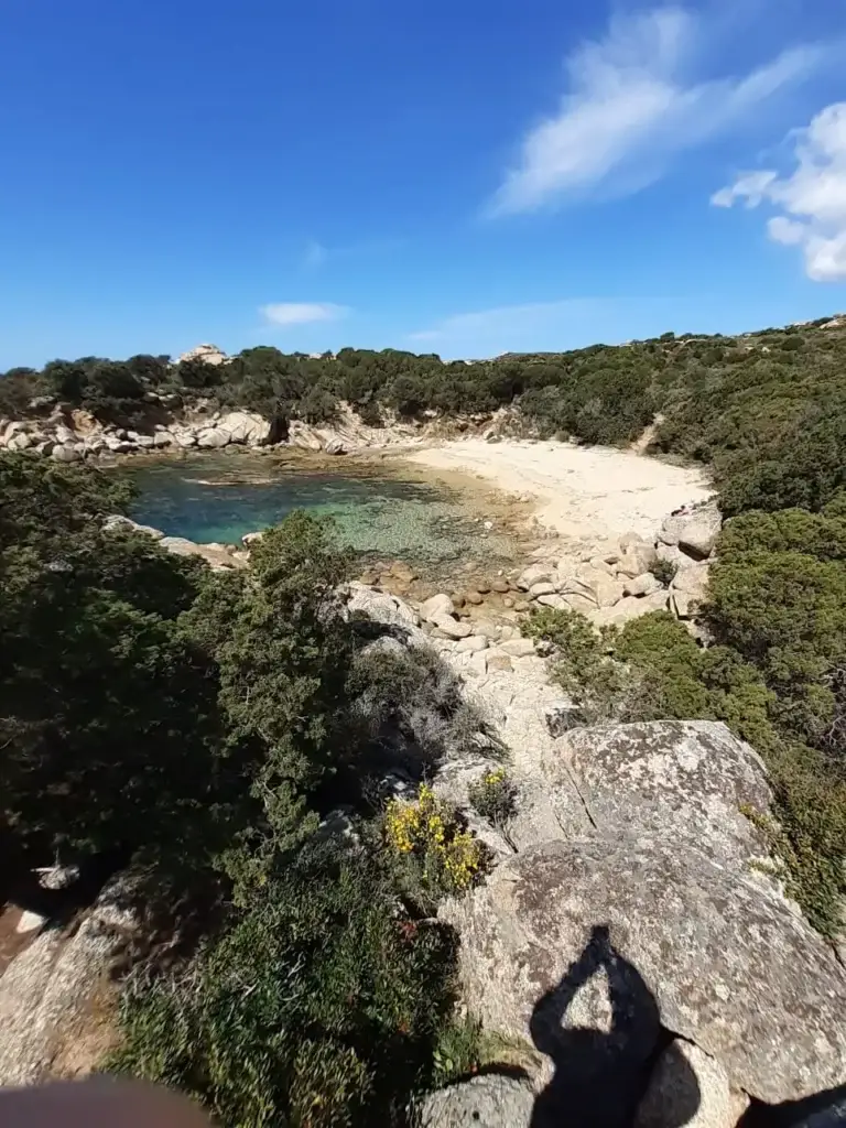 Vue d'en haut sur une baie rocheuse abritée avec une plage de galets en Corse. Les eaux sont transparentes et la végétation de maquis environnante est dense. L'ombre de la personne qui prend la photo est projetée sur un rocher au premier plan.